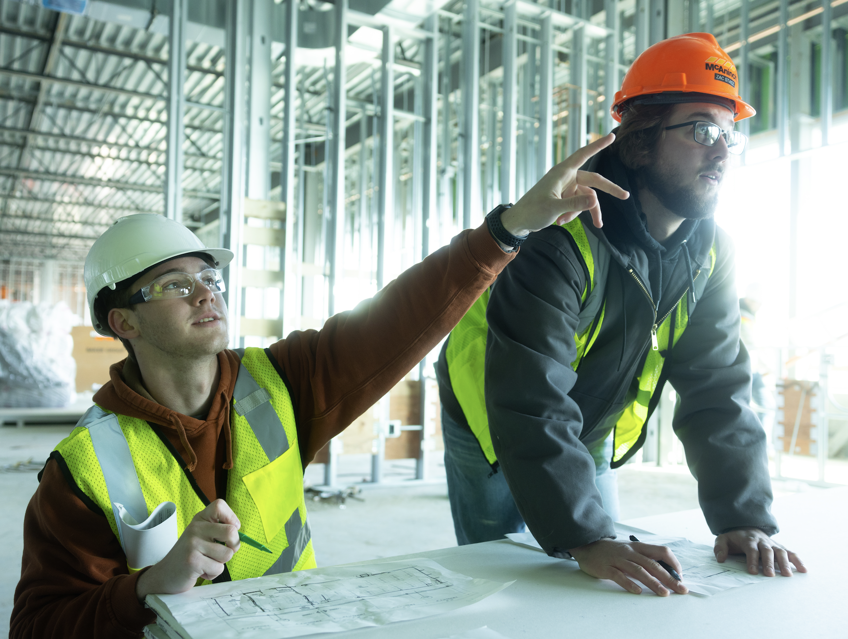 Construction engineering students visiting a job site wearing hard hats, protective eyewear and neon vests. The student on the left points off-camera.